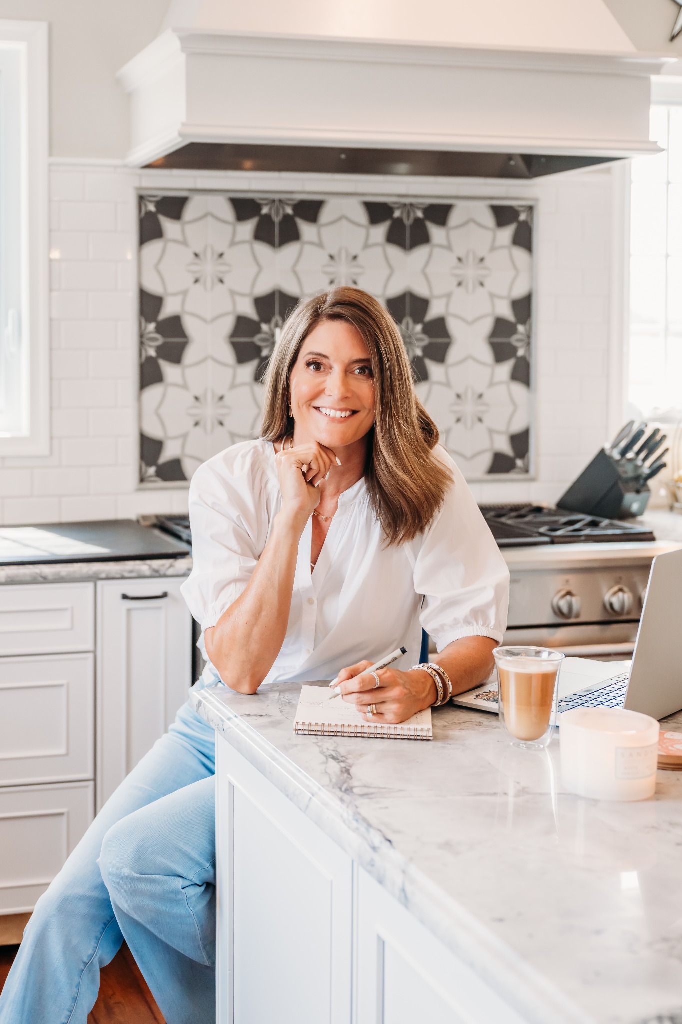 Mandy Corcoran, founder of Creative Systems Lab, smiling at the camera in her kitchen with her own pattern design as a tile backsplash behind her